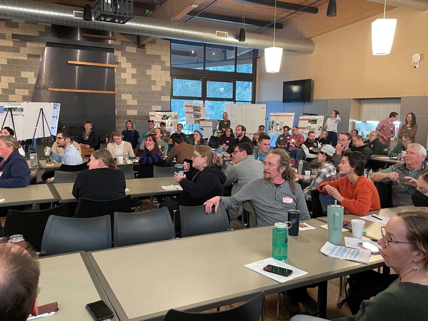 Picture4 a photo of a large group of people in an assembly hall watching a presentation. There are scientific posters displayed in the background.